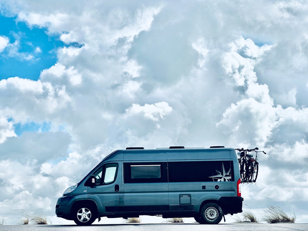 A blue van parked under a picturesque cloudy sky in Dunkirk, France, with a bicycle attached to the back.