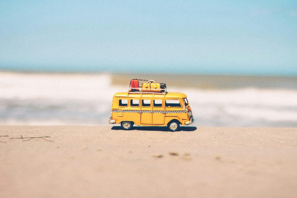 A vibrant yellow toy van sits on a sandy beach with an ocean backdrop, symbolizing fun travel adventures.