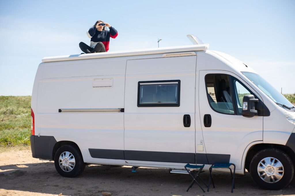 A woman sitting on the roof of a camper van using binoculars during a sunny day.